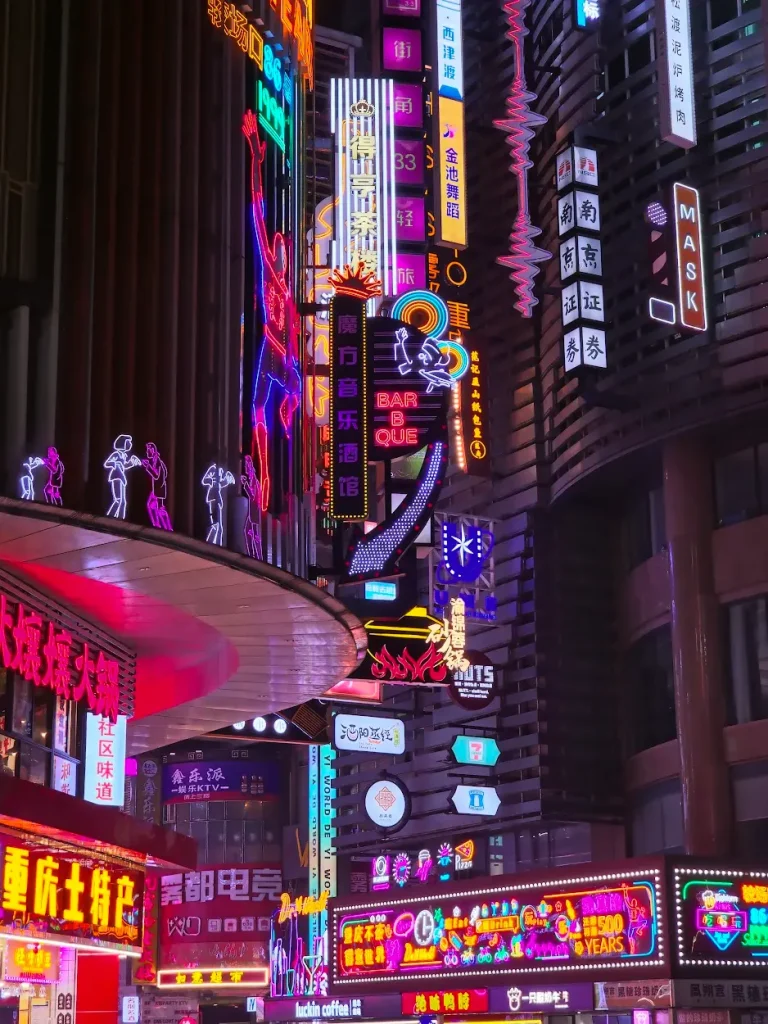 Colorful neon signs and billboards in various languages illuminating a busy urban street at night.