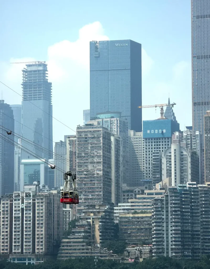 Red cable car suspended over a densely packed cityscape with tall modern and older high-rise buildings.